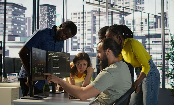 A diverse group of employees gathered around a computer screen discussing deployment rules and company policy changes.