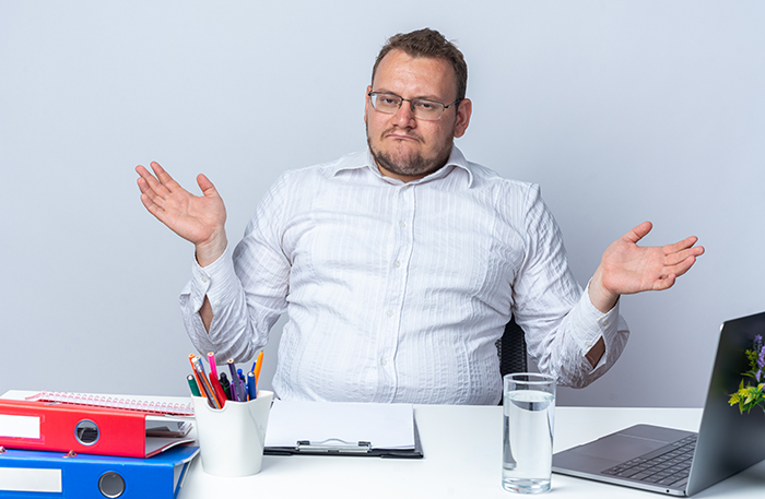 Man sitting at desk with laptop and files, shrugging as he adjusts to stick to his schedule after being told to stop being late.