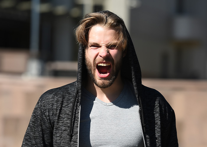 Man wearing a hoodie yelling angrily outdoors, expressing frustration related to washing and waxing a car after egg throwing.