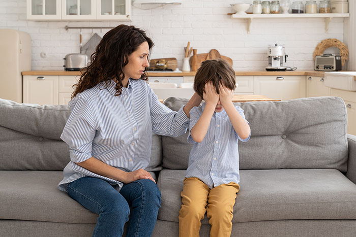 Woman babysitting nephew for SIL looks concerned as young boy covers his face on a living room couch. Woman babysitting nephew for SIL looks concerned as young boy covers his face on a living room couch.