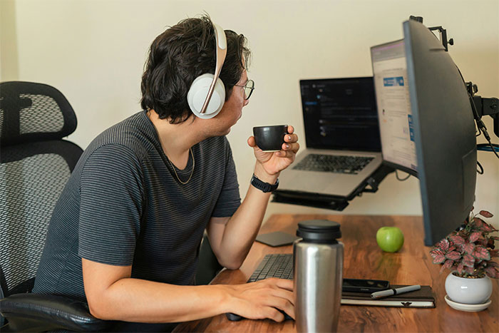 Employee following unworkable deployment rule wearing headphones, working at dual monitors, and holding a cup in office setting.