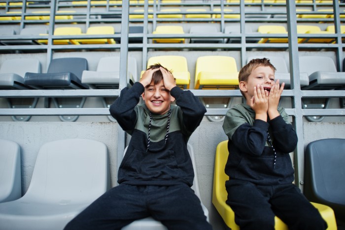 Two boys in matching outfits sitting on stadium seats, one laughing and holding his head, the other covering his mouth.