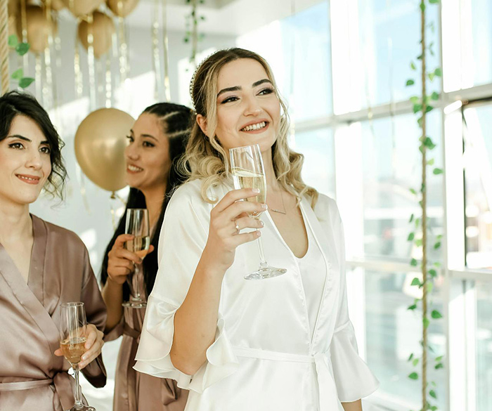 Group of women in robes celebrating with champagne glasses at a bachelorette party indoors with decorations.