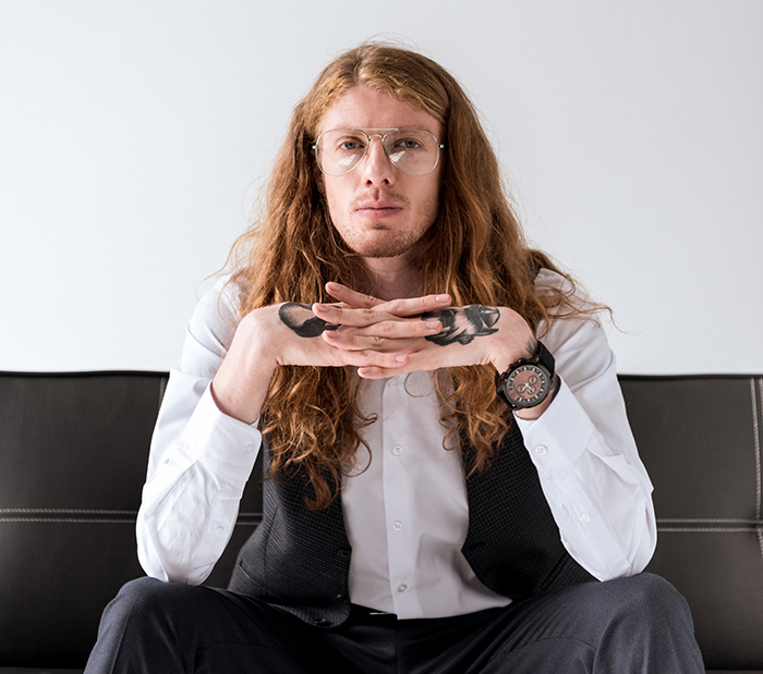 Young man with long hair and tattoos on hands, wearing glasses and a formal vest, representing good and bad genes.