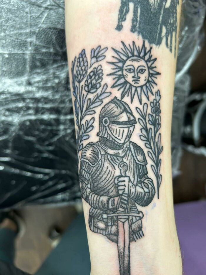 Tattoo of a knight in armor holding a sword with floral elements and a sun face, a bad tattoo inkshaming example.