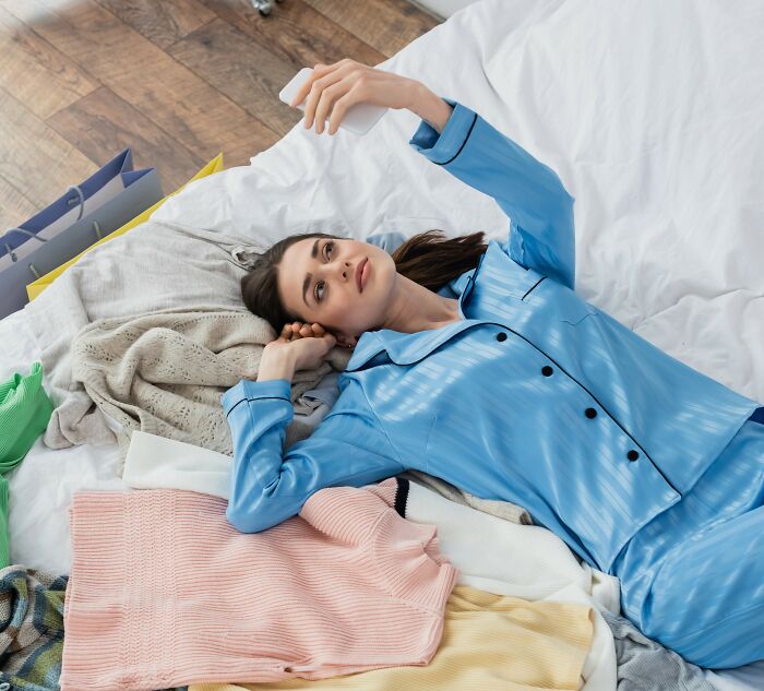 Woman over 42 in blue pajamas lying on bed surrounded by clothes, using smartphone and reflecting on what she won’t tolerate.