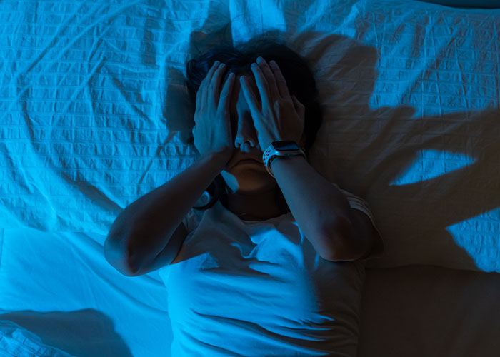 Person lying in bed covering face with hands, illustrating creepy facts that might get stuck in your brain forever concept.