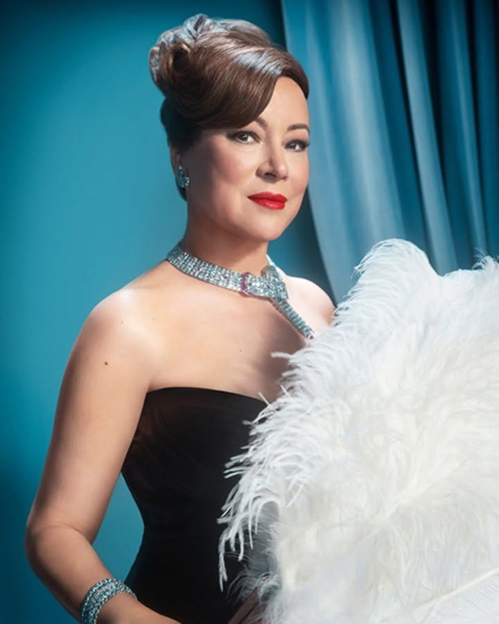 Jennifer Tilly dressed glamorously with elegant jewelry, posing against a teal background holding white feathers.