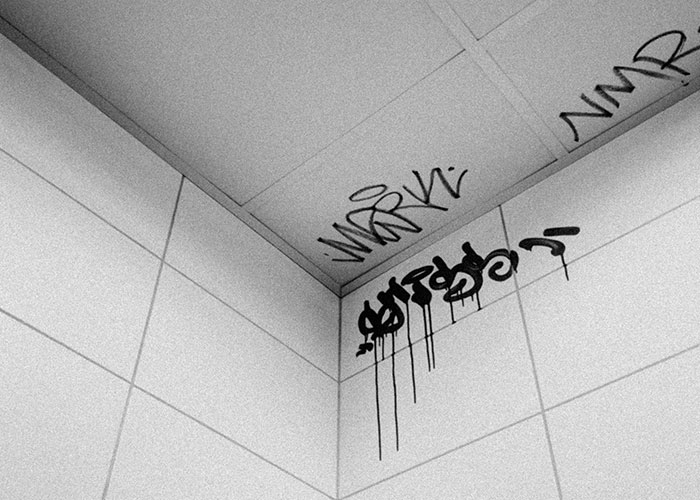 Black and white photo of graffiti on tiled walls and ceiling corner, depicting a raw urban take one for the team moment.