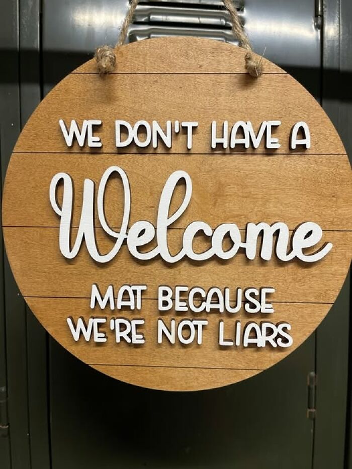 Funny chaotic sign on wooden board saying no welcome mat because we're not liars, adding humor and chaos to the world.