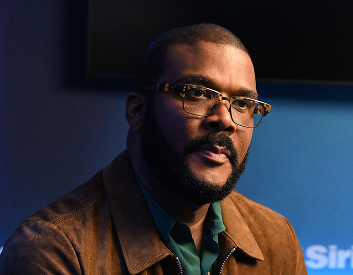 Tyler Perry wearing glasses and a brown jacket in a serious moment related to harassment allegations discussion.