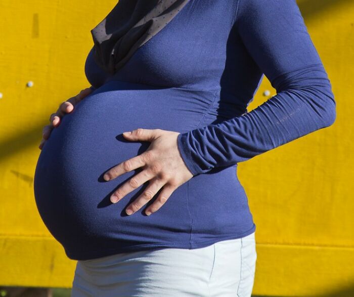 Pregnant woman in blue shirt gently holding baby bump against a bright yellow background, illustrating twins pregnancy.