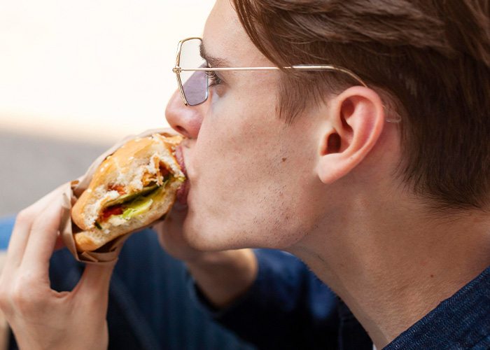 Young man wearing glasses eating a sandwich, capturing a moment on public transport with a humorous vibe