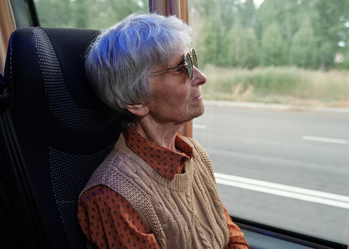 Elderly woman wearing sunglasses looking out the window of a public transport bus on a rural road.