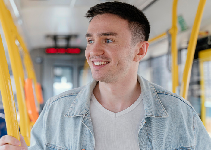 A young man smiling inside a public transport vehicle, holding a yellow handrail and looking out the window.