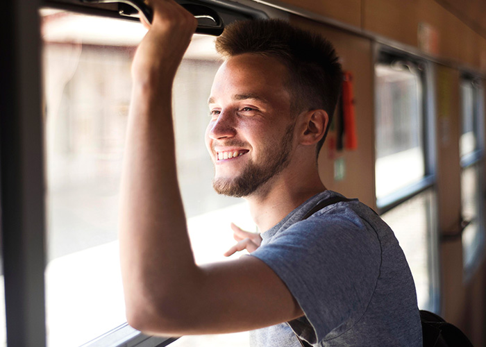 Smiling young man standing and holding a strap inside public transport, enjoying his ride on a sunny day.