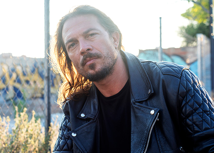 Man with long hair and beard wearing a black leather jacket, captured in natural light with a blurred outdoor background.