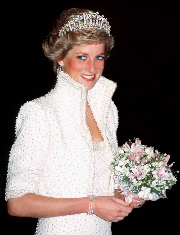 Princess Diana wearing a pearl-embellished white dress and tiara, smiling while holding a bouquet of flowers, showcasing famous good looking names.