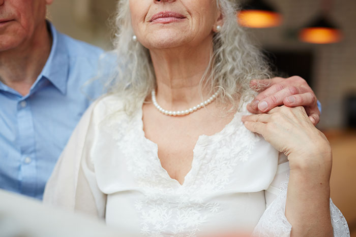 Groom&rsquo;s mom in a wedding dress with pearl necklace, holding a man's hand, attending a wedding event indoors.