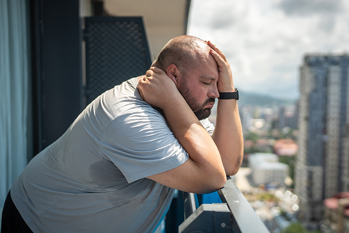 Man experiencing bil struggles on balcony during China trip, holding head and neck in visible discomfort.