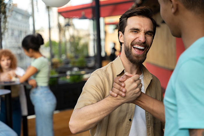 Two men shaking hands and laughing at an outdoor cafe, capturing a moment of friendly interaction and banter. Two men shaking hands and laughing at an outdoor cafe, capturing a moment of friendly interaction and banter.