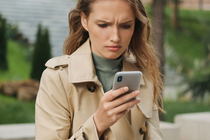 Young woman in beige coat looking upset while checking her phone, related to refusing to pay cat-sitter issue.
