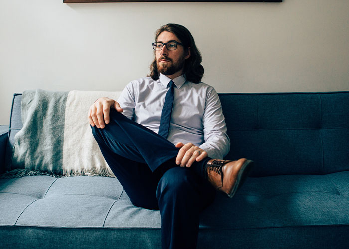 Man with glasses and long hair sitting thoughtfully on a blue couch, evoking creepy facts stuck in your brain.