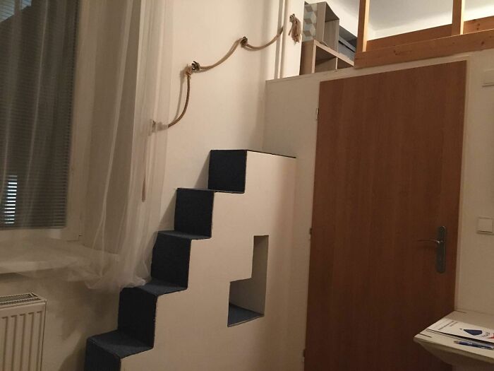 Narrow and steep death stairs with a small opening, featuring a rope handrail in a dimly lit room next to a wooden door.