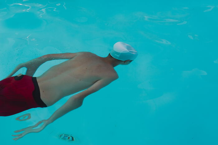 A swimmer wearing a white cap and red shorts diving underwater, captured in a perfectly-timed street photo by Luca Regoli.