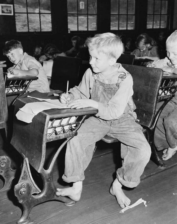 Barefoot young boy in overalls writing at a vintage wooden school desk in a historical classroom setting.