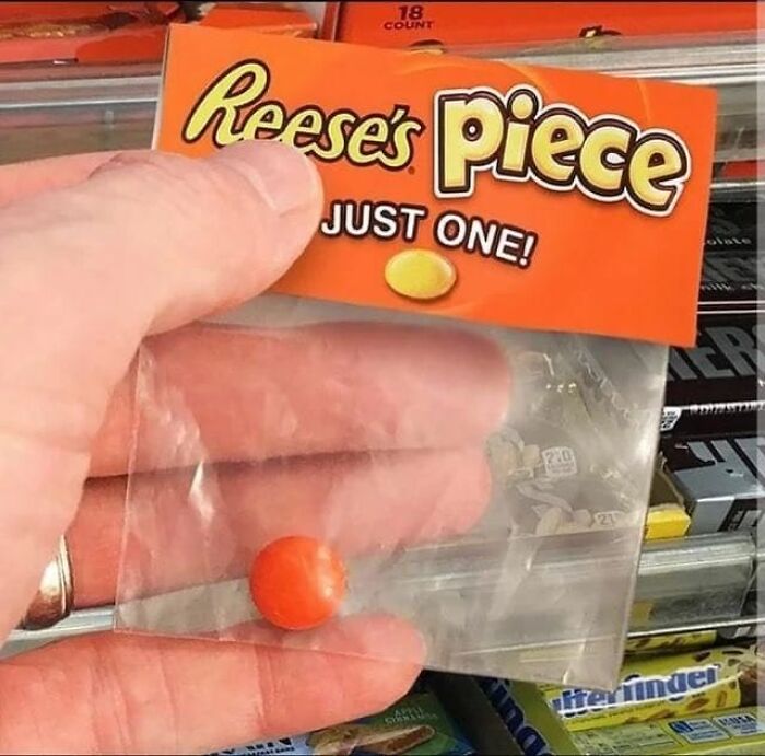 Hand holding a single Reese's Piece candy in a mostly empty small plastic bag in a store display, cursed food image.