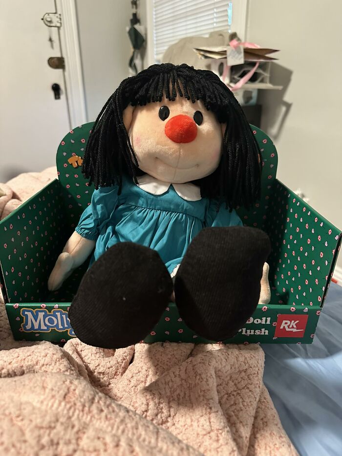Plush doll with black yarn hair and red nose in green box, representing nostalgia for people between Millennials and Gen Z.