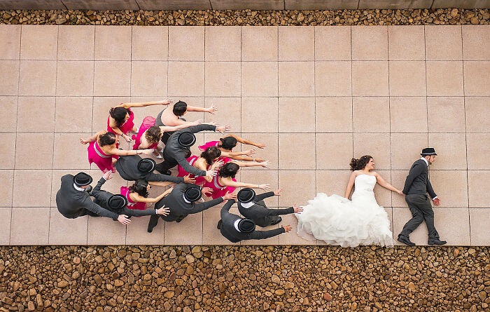 Overhead view of a bride in white gown and groom walking away while bridesmaids and groomsmen reach out in wedding photo.