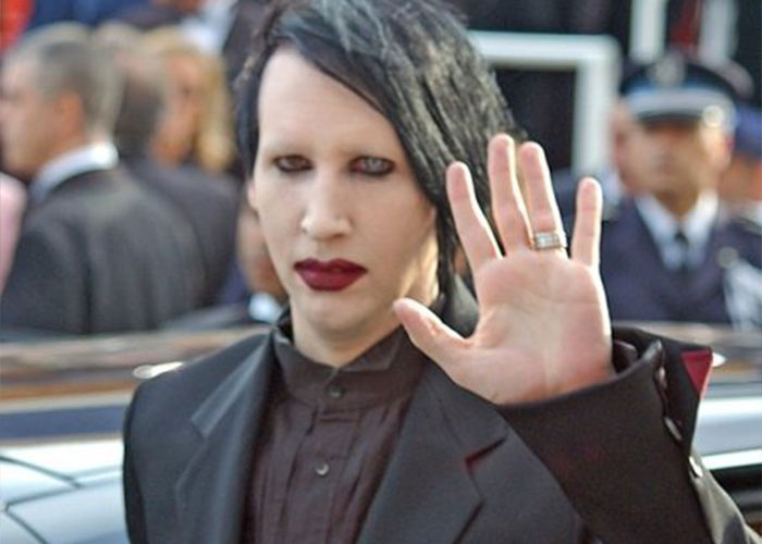 Person with heavy makeup and dark clothing raising a hand, representing unhinged men's comments about women's looks in public.