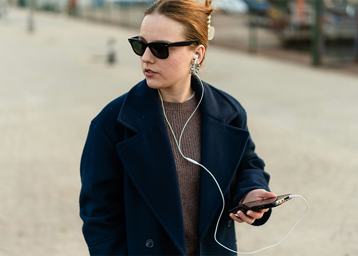 Young woman wearing sunglasses and earbuds, holding a smartphone while embracing obsolete technology use outdoors.