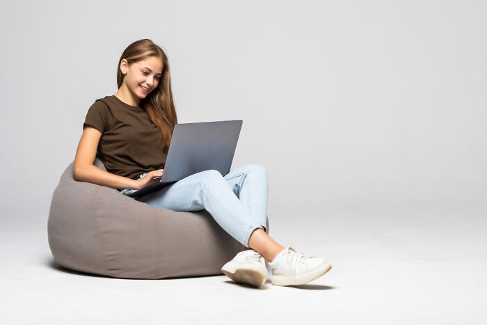 Young woman using a laptop while sitting comfortably on a bean bag, representing out-of-touch rich people flaunting wealth.