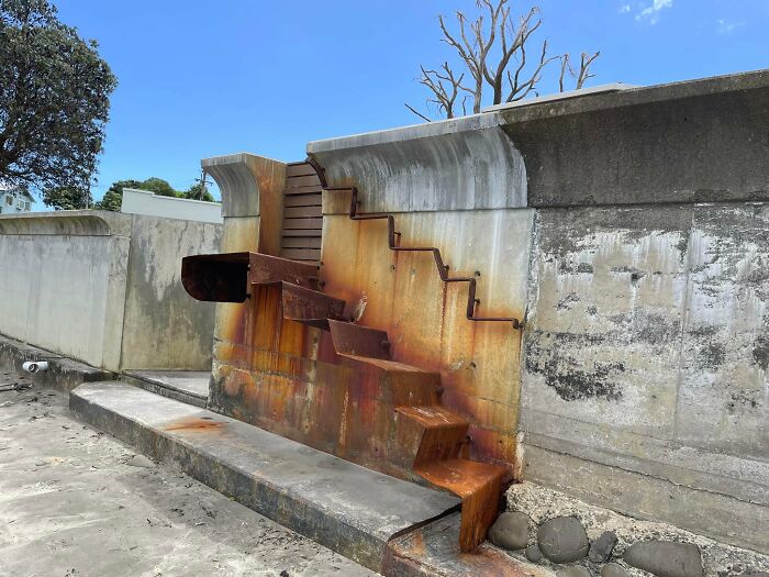Rusty death stairs with irregular metal steps attached to a concrete wall next to a sandy beach under clear blue sky.