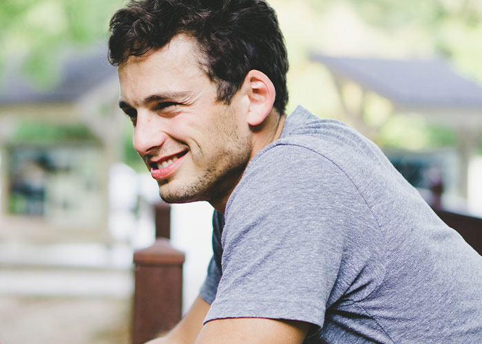 Young man in a grey shirt smiling while sitting outdoors, illustrating unhinged things men have said about women's looks in public.