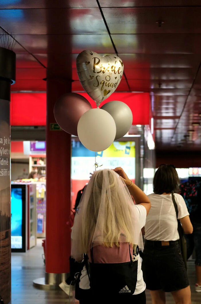 Bride squad balloons held by a woman in a veil walking in a busy indoor public space, street photography capturing everyday life.