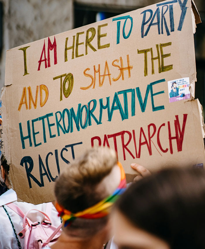 Protest sign about smashing heteronormative racist patriarchy held by a millennial at a crowded event.