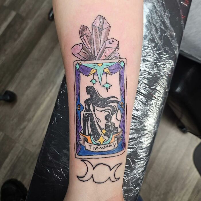 Tattoo of a crystal cluster with a framed figure and text, accompanied by a triple moon symbol on the forearm in bright colors.