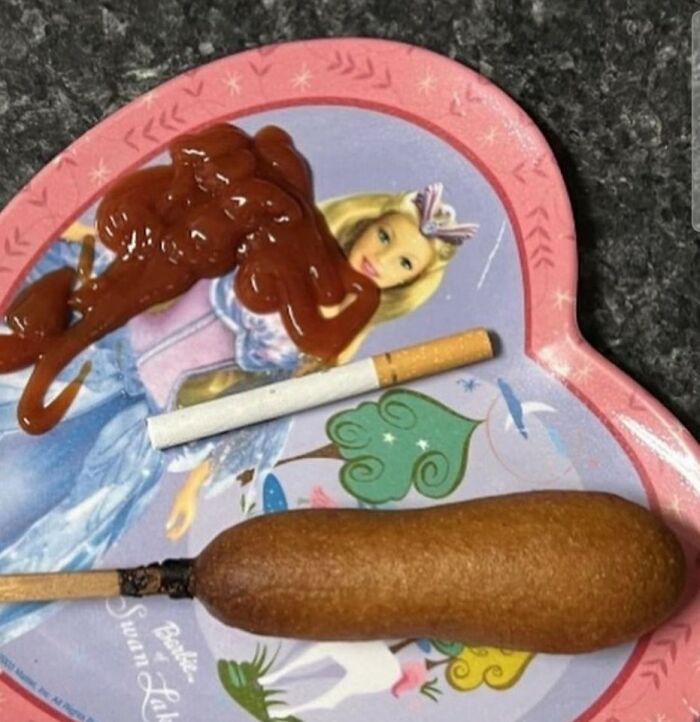 Cursed food image with ketchup on princess plate, cigarette, and burnt corn dog on dark countertop.