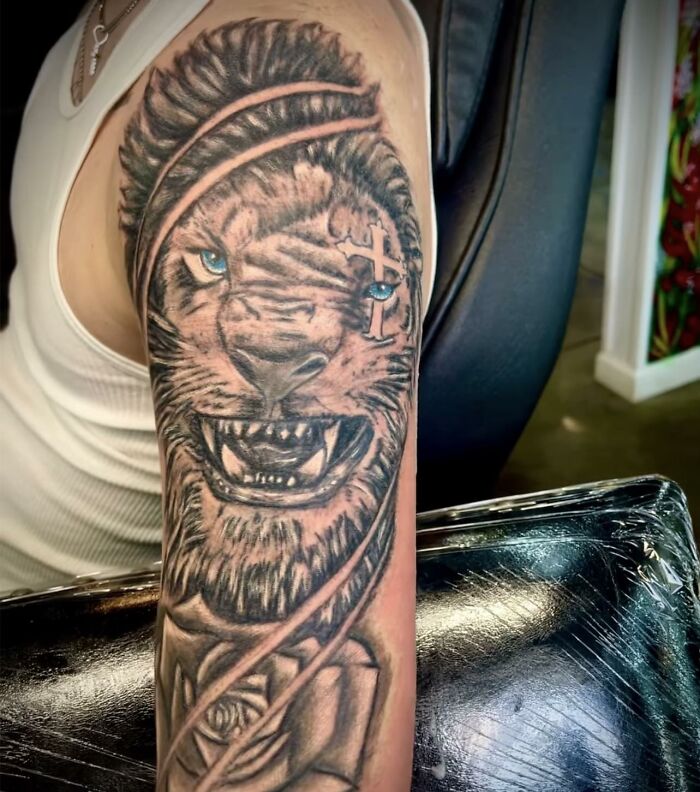 Lion tattoo with blue eyes and a cross design on a person's upper arm, an example of hilariously bad tattoos.