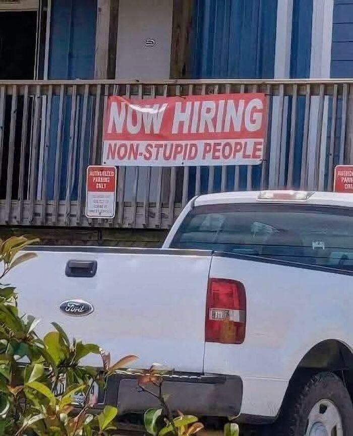 Funny sign reading now hiring non-stupid people posted on blue porch railing behind white Ford pickup truck.