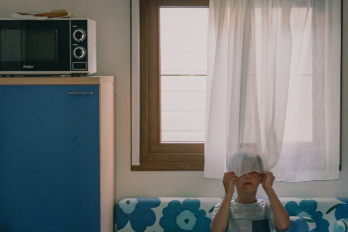 Child playfully hiding behind a curtain in a kitchen, captured in a perfectly-timed street photo by Luca Regoli.