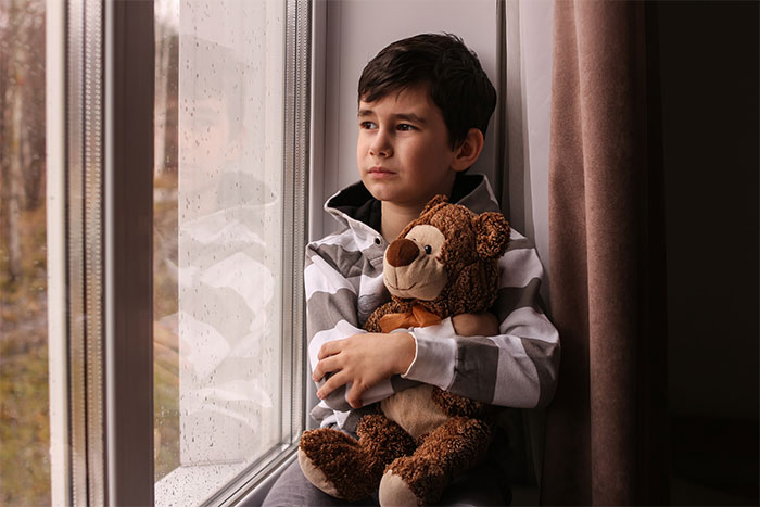 Sad young boy clutching teddy bear by window, reflecting feelings of neglect and emotional struggle with parents.