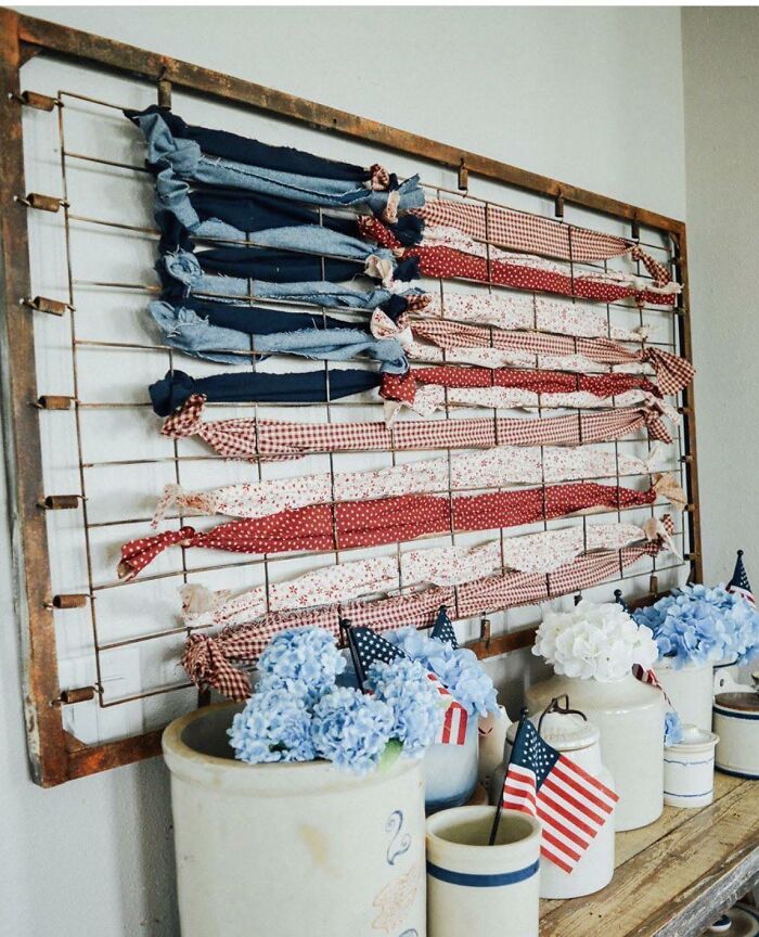 Rustic American flag craft made with fabric strips on a metal frame above vintage jars with blue and white flowers.
