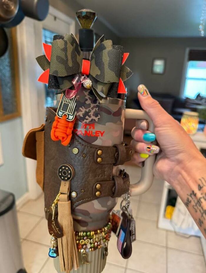 Hand holding a camo Stanley mug covered in an unusual craft mix of leather, beads, bows, and charms, showcasing a horrible craft fail.