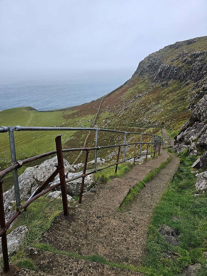 Steep and narrow death stairs with rusty railing descending a cliffside path overlooking the ocean on a cloudy day.