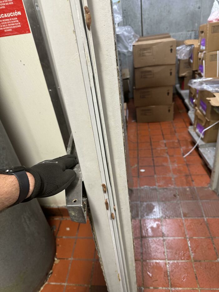 Hand wearing a black glove opening a door to a storage room with cardboard boxes on a red tiled floor, showing signs of neglect.